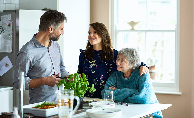 young woman with her arm around older woman while talking with a man in the kitchen