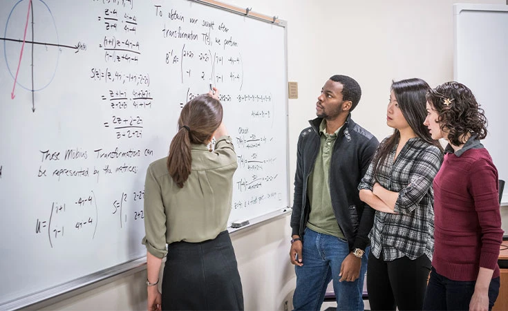 female teacher pointing to a math problem on a white board and explaining it to three adult students