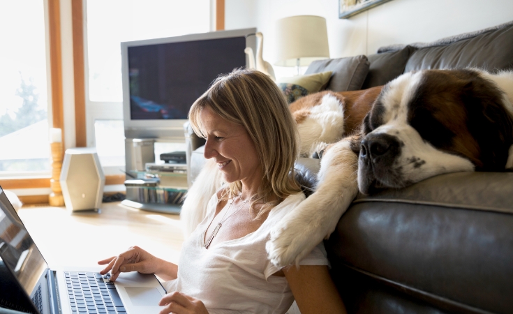 young white woman smiling while sitting with her dog and looking at her laptop