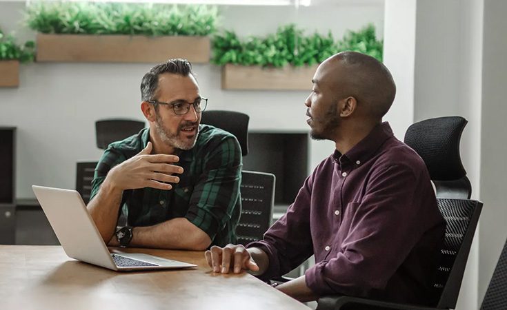 two men sitting at a conference table with a laptop and talking
