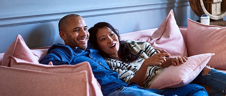 Mature man with arm around Vietnamese woman, sitting with feet up and smiling, woman pointing to TV, entertainment, relaxation, togetherness