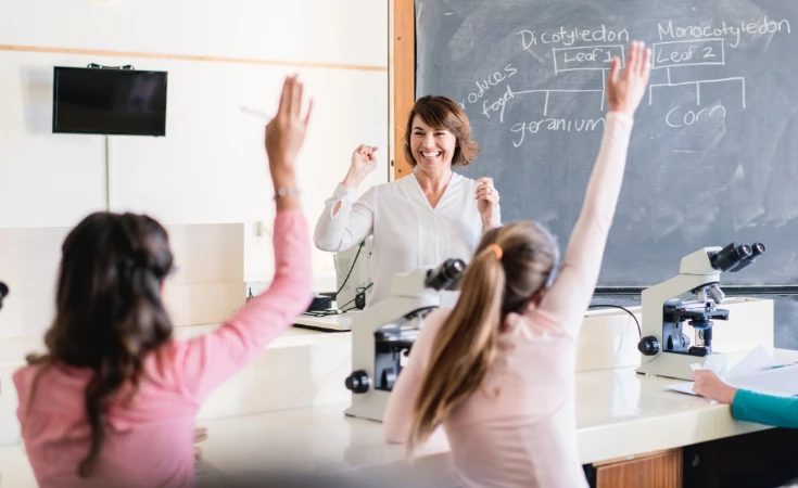Teacher smiling at students who are raising their hands in a science classroom with microscopes 