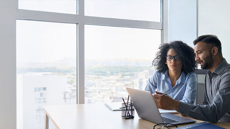 Two coworkers sit together at a desk, collaborating while looking at a laptop screen