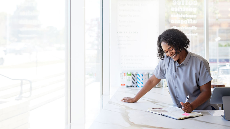 A person stands at a modern reception counter, writing in a notebook with a pen
