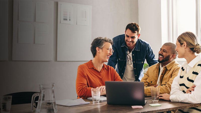 A small group of colleagues gathers around a laptop at a wooden table in a modern office